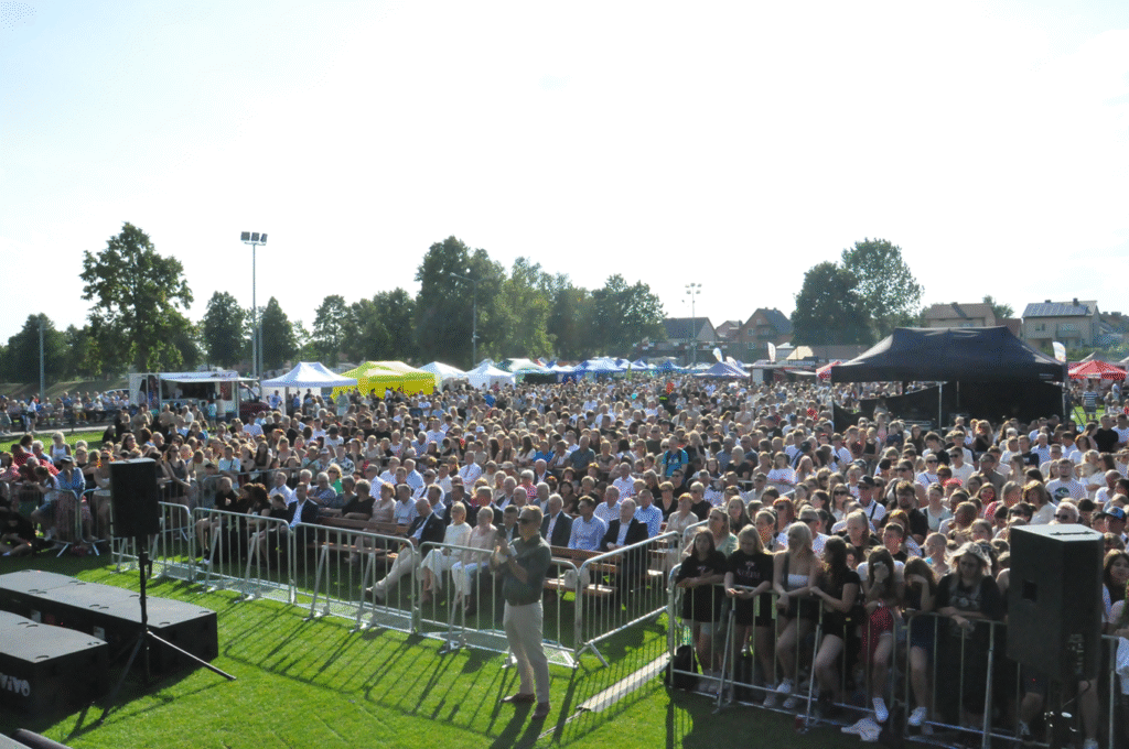 Tłum ludzi. W tle namioty z atrakcjami, budynki mieszkalne i drzewa. Słoneczny dzień.