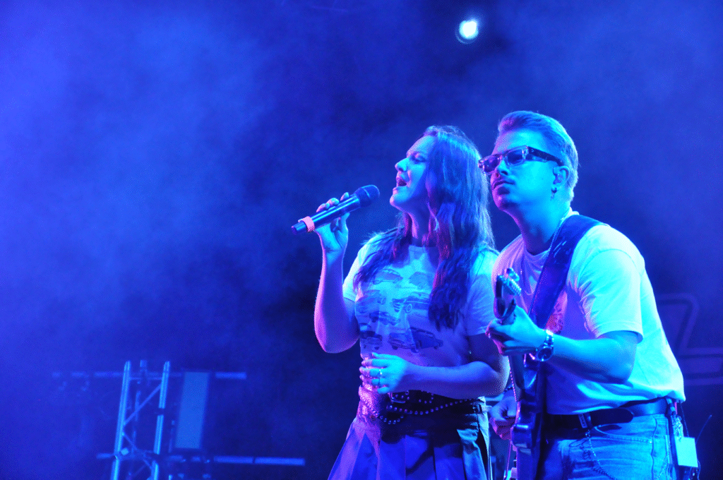 Młoda kobieta w białej koszulce z nadrukiem samochodów trzyma mikrofon przy ustach. Obok niej stoi mężczyzna z gitarą elektryczną. Wieczorna scena plenerowa.