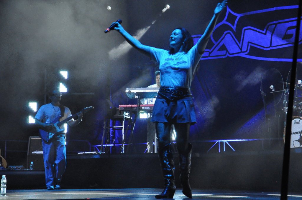 Kobiet w białej koszulce z nadrukiem samochodów wznosi ręce. Za nią gitarzysta i perkusista. Wieczorna scena plenerowa.