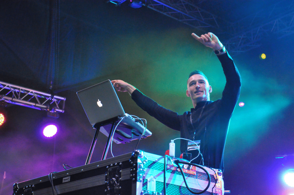 Mężczyzna w czarnej koszuli stoi przy laptopie i wymachuje rękami. Wieczorna scena plenerowa.