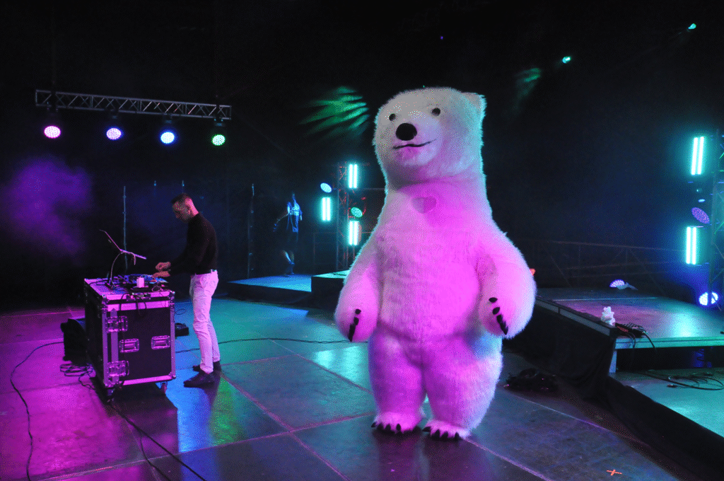 Dj w czarnej koszuli i w jasnych spodniach przy sprzęcie muzyczny. Obok niego osoba przebrana za niedźwiedzia polarnego. Wieczorna scena plenerowa.