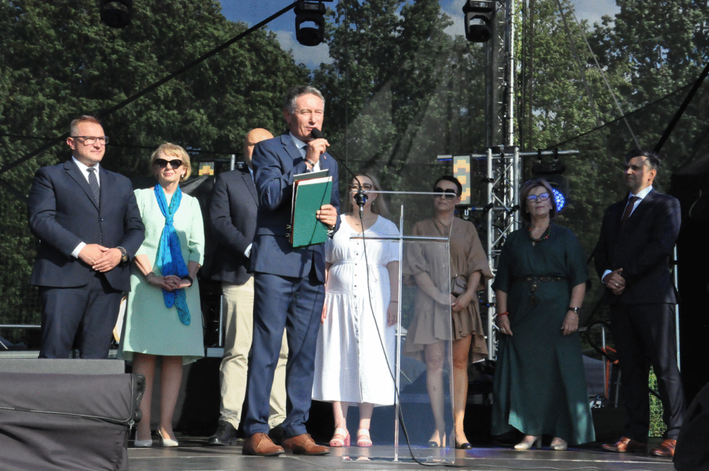 Mężczyzna w grafitowym garniturze przemawia do mikrofonu na statywie . Za nimi grupa stojących osób, kobiety i mężczyźni w oficjalnych strojach. W tle korony drzew.