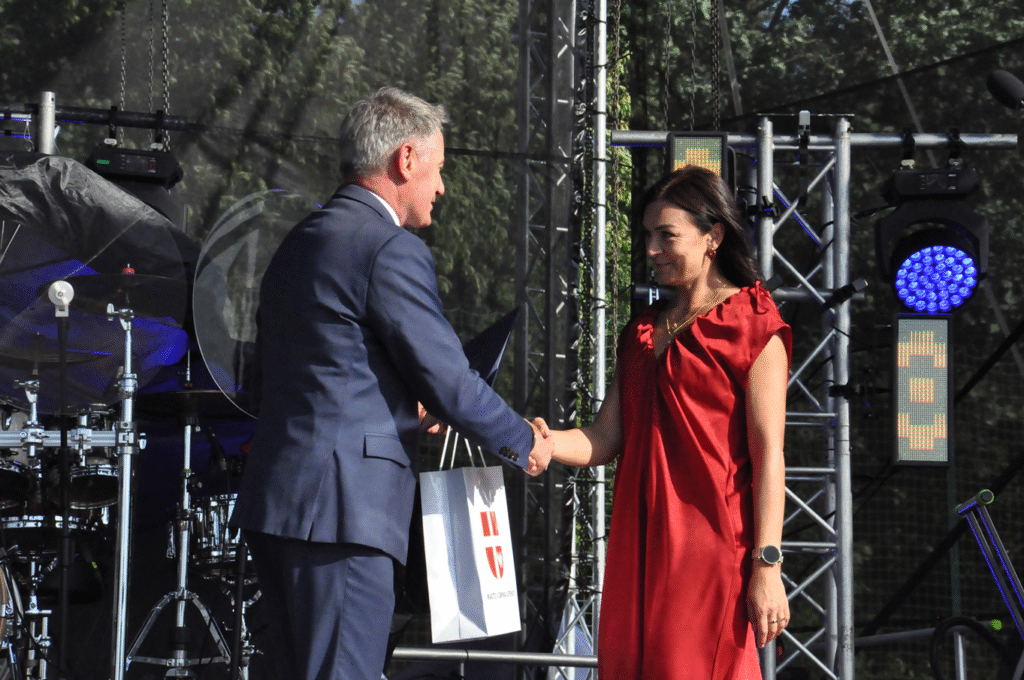 Mężczyzna w grafitowym garniturze podaje dłoń kobiecie w czerwonej sukience. W tle sprzęty elektroniczne.