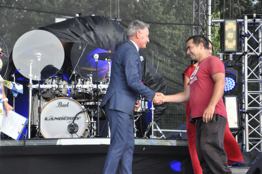 Mężczyzna w grafitowym garniturze podaje dłoń mężczyźnie w czerwonej koszulce z białym napisem. W tle instrumenty muzyczne i sprzęty elektrorniczne.