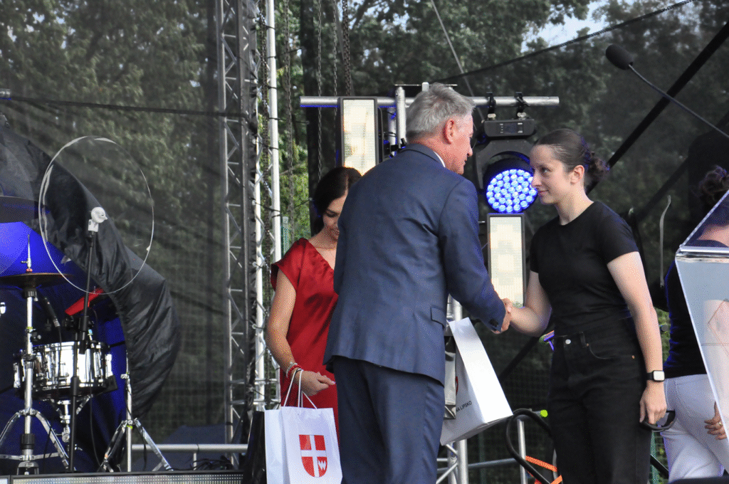 Mężczyzna w grafitowym garniturze podaje dłoń dziewczynie w czarnej koszulce i czarnych spodniach. Za nimi stoi kobieta w czerwonej sukience. W tle instrumenty muzyczne i sprzęty elektrorniczn.