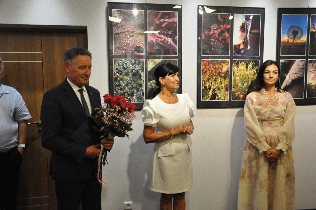 Mężczyzna w grafitowej marynarce trzyma bukiet czerwonych kwiatów. Obok niego stoją dwie kobiety w jasnych sukienkach. Za nimi fotografie w antyramach.