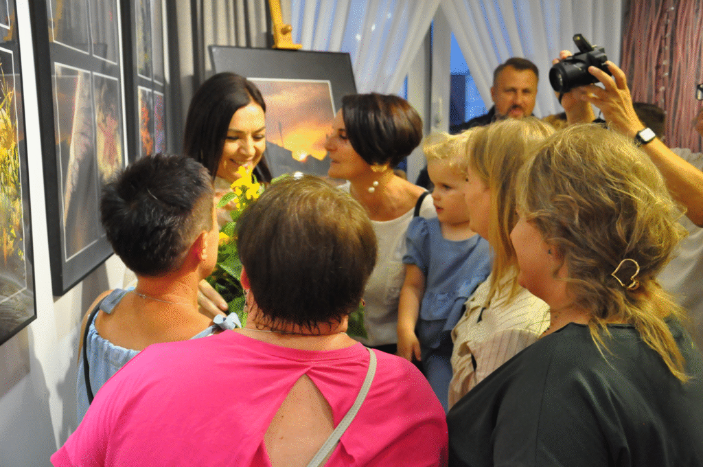 Kilka osób zgromadzonych przy artystce. Pomieszczenie ciepło oświetlone. Na ścianie antyramy z fotografiami.