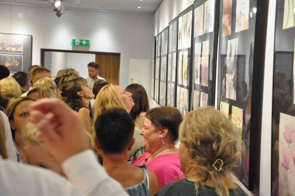 Ludzie przyglądają się ścianie na której wiszą antyramy z pracami malarskimi. Pomieszczenie ciepło oświetlone.