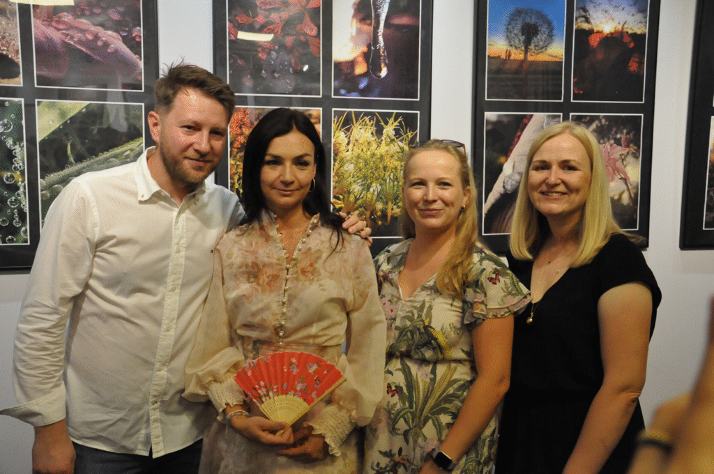 Trzy kobiety i mężczyzna pozują do pamiątkowego zdjęcia. Jedna kobieta trzyma czerwony wachlarzyk. Za nimi wiszą antyramy z fotografiami.