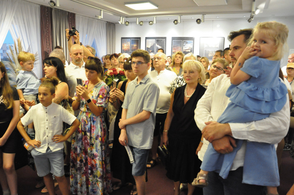 Zgromadzeni ludzie stoją w ciepło oświetlonym pomieszczeniu. W tle ściana z fotografiami w antyramach.