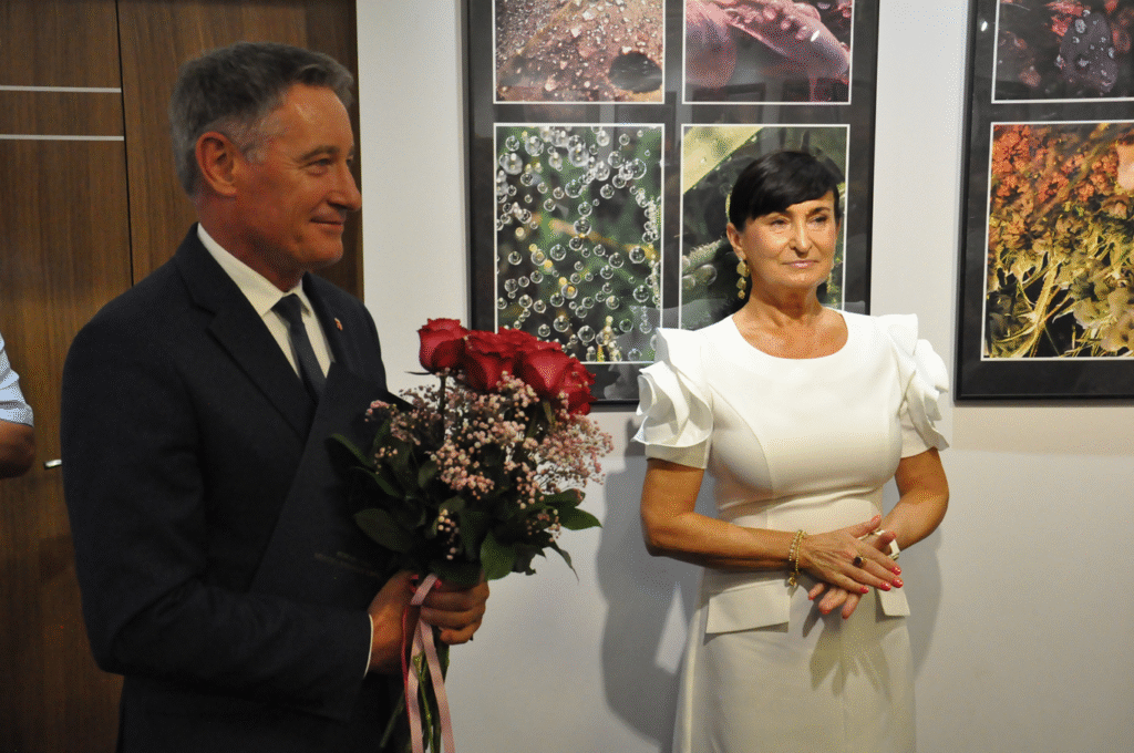 Mężczyzna w grafitowej marynarce trzyma bukiet czerwonych kwiatów. Obok niego stoi kobieta w jasnej sukience. Za nimi fotografie w antyramach.