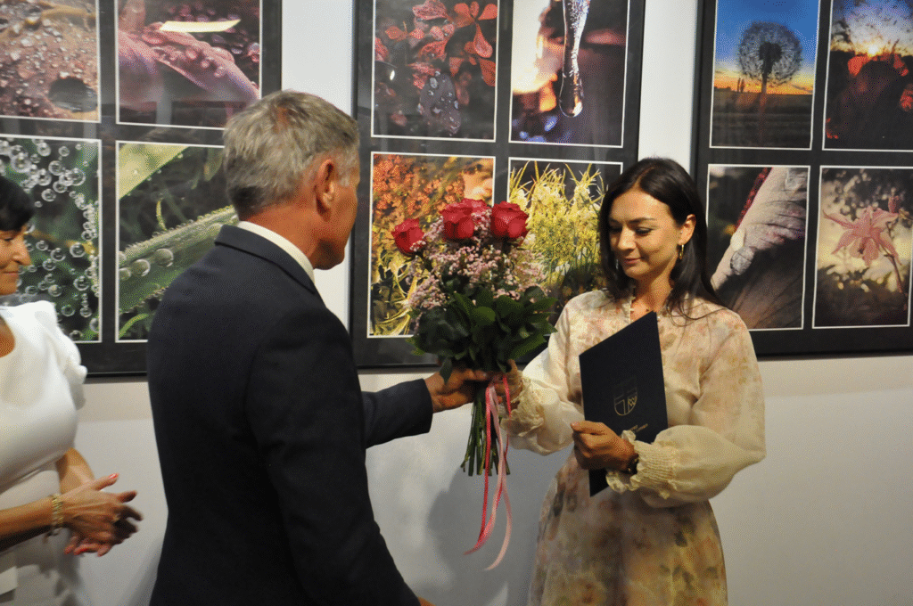 Mężczyzna w grafitowej marynarce podaje bukiet czerwonych kwiatów kobiecie w jasnej sukienkce. Za nimi fotografie w antyramach.