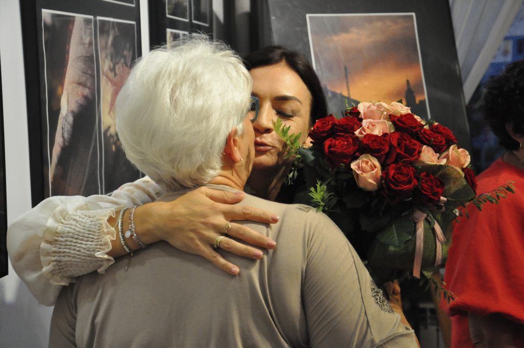 Kobieta z bukietem czerwonych kwiatów całuje w policzek kobietę w szarej bluzce. Za nimi wiszą antyramy z fotografiami.