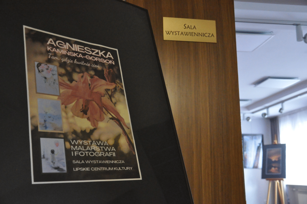Plakat z informacjami o wystawie w czarnej antyramie na drugim planie drzwi z tabliczką "sala wystawiennicza", jeszcze dalej wnętrze sali wystawienniczej.