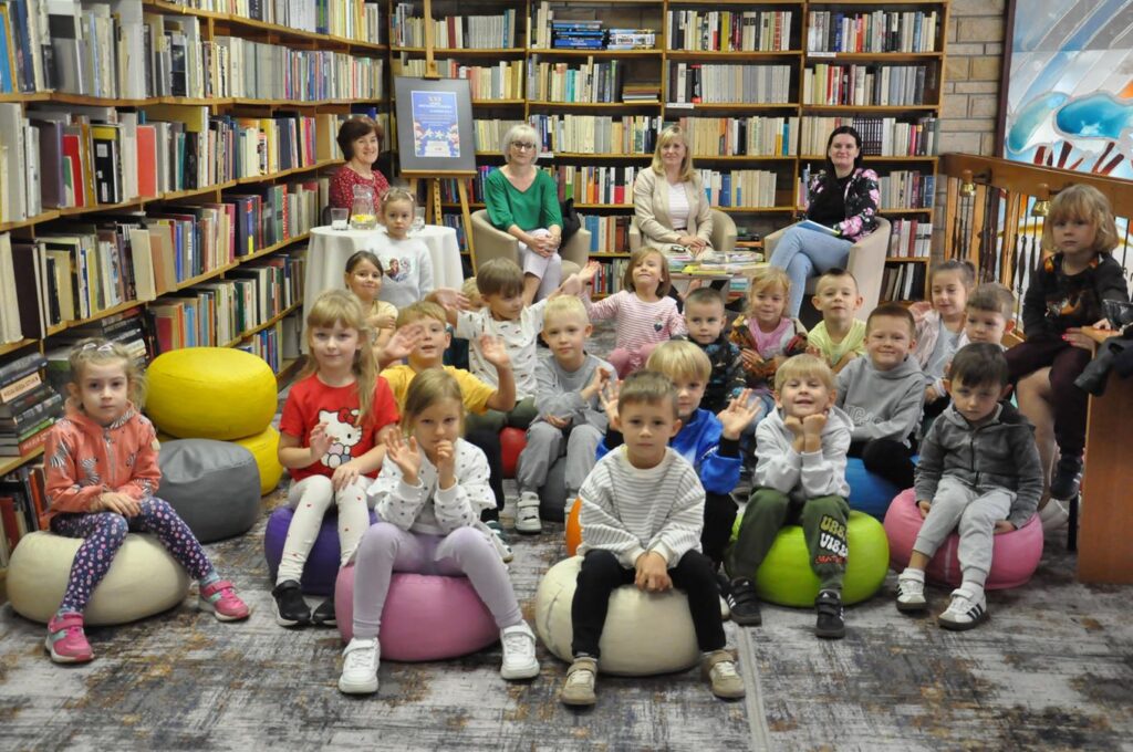 Cztery kobiety i grupa dzieci. Pozują do wspólnego zdjęcia. Wszyscy siedzą. Wokół regały z książkami.
