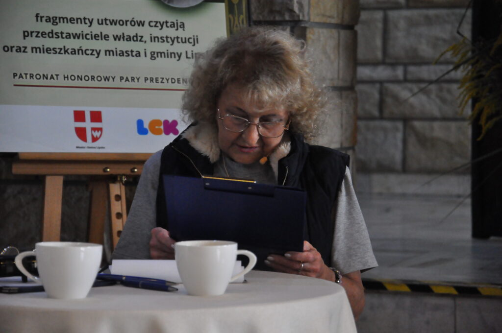 Kobieta z siwymi włosami, okularami i czarnej kamizelce siedzi przy stole na którym leżą filiżanki. Trzyma granatową podkładkę na dokumenty.
