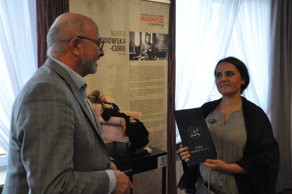 Kobieta w szaro-czarnej koszuli trzyma tekturową, granatową teczkę z logotypem jednostki kultury. Obok niej stoi mężczyzna w szarej marynarce i okularach. Za nimi rollup wystawy.