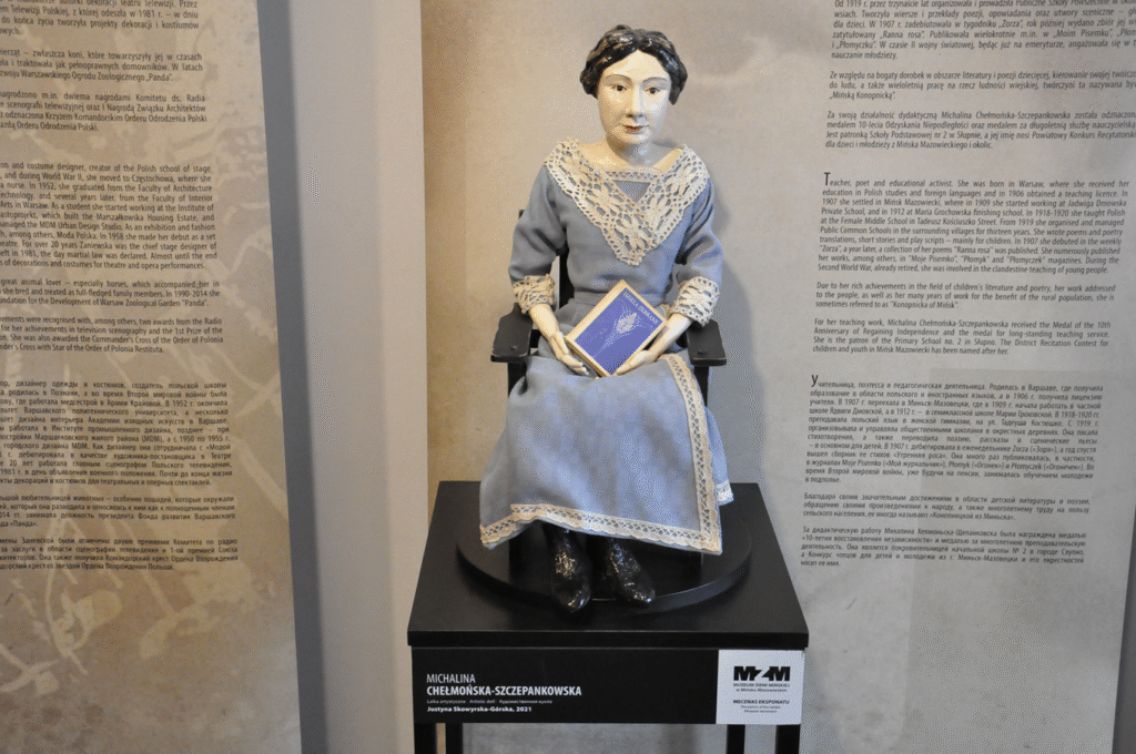 Ekspozycja porcelanowej lalki w błękitnej długiej sukni z książką na kolanach, która siedzi na krześle.