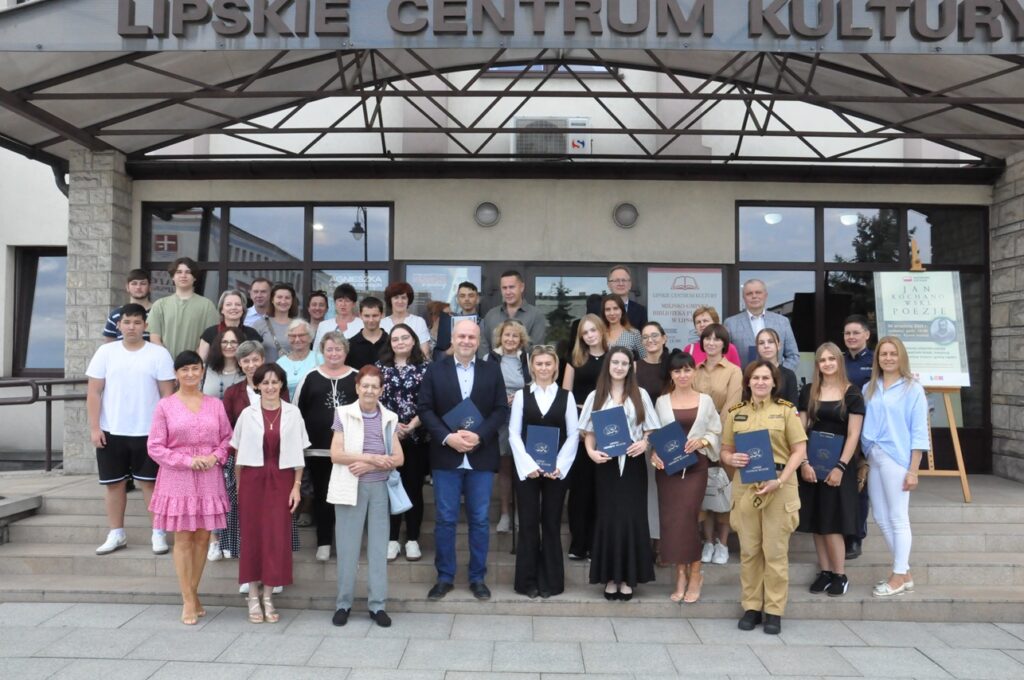 Duża grupa ludzi stoi na schodach przed wejściem do jednostki kultury. Niektórzy trzymają papierowe granatowe teczki na dokumenty. Po prawej stornie stoi stelaż z plakatem promującym wydarzenie.