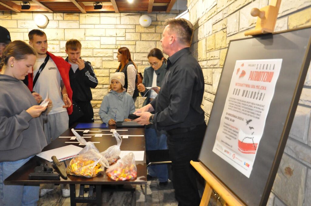 Mężczyzna w szarej koszuli stoi obok stelaża z plakatem obok stołu na którym leżą woreczki z cukierkami i kartki papieru. Po drugiej stronie stołu stoją młodzi ludzie.
