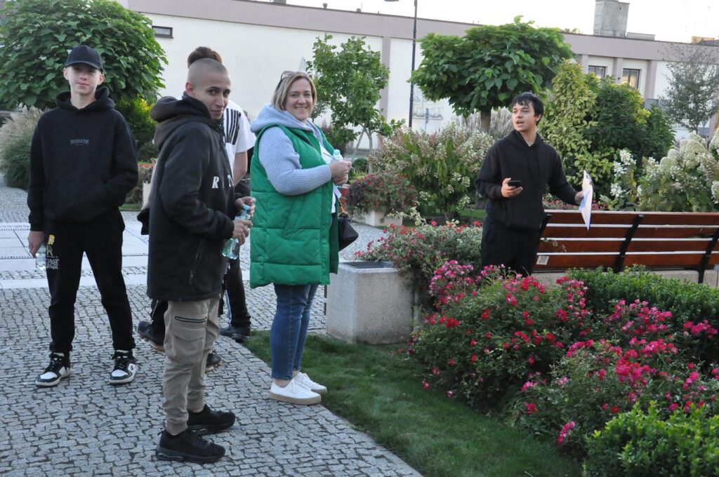 Grupa młodzieży i kobieta w zielonej kurtce stoi w miejskim parku.