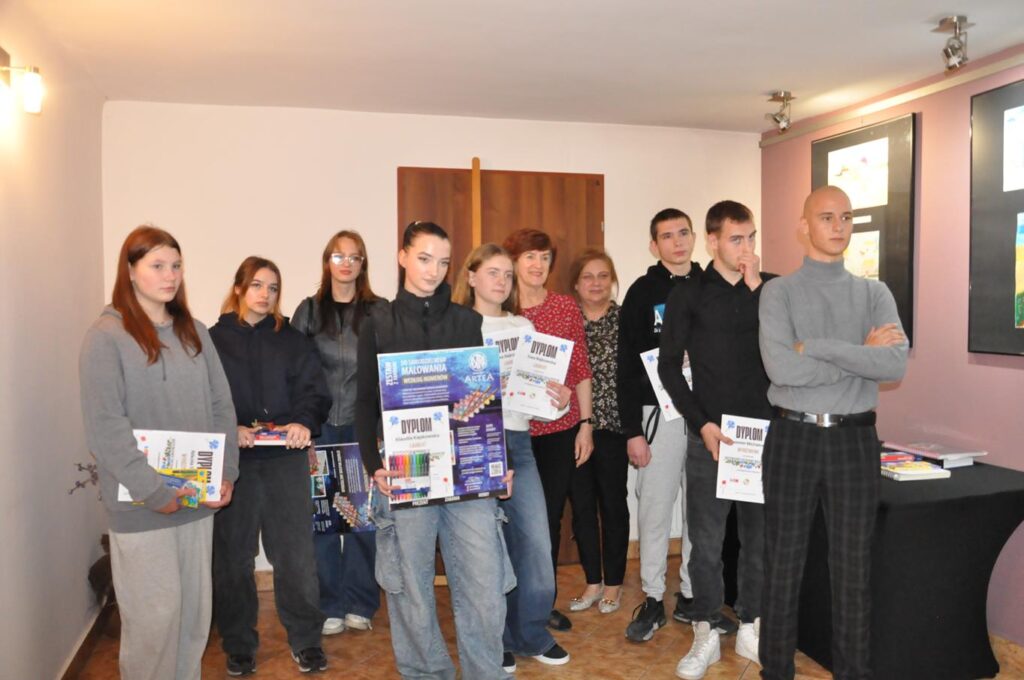 Grupa młodzieży pozuje do wspólnego zdjęcia. Niektórzy trzymają dyplomy i nagrody. Pomieszczenie ciepło oświetlone.