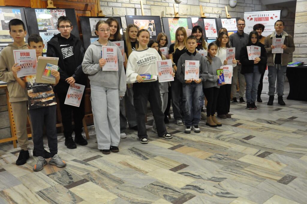 Uczestnicy konkursu pozują do wspólnego zdjęcia i eksponują pamiątkowe dyplomy i nagrody. Pomieszczenie ciepło oświetlone. Za nimi ekspozycja plastycznych prac.
