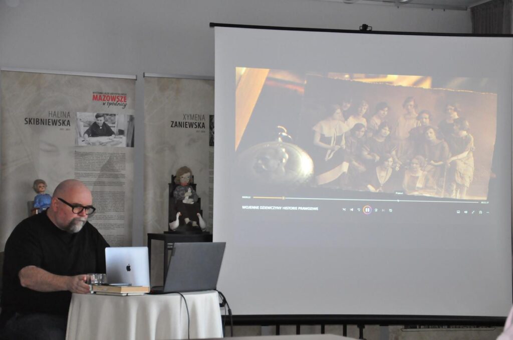 Mężczyzna z siwą bródką i czarnych okularach siedzi przy okrągłym stole na którym stoją dwa laptopy. Obok niego duży ekran z wyświetlonym kadrem filmu.