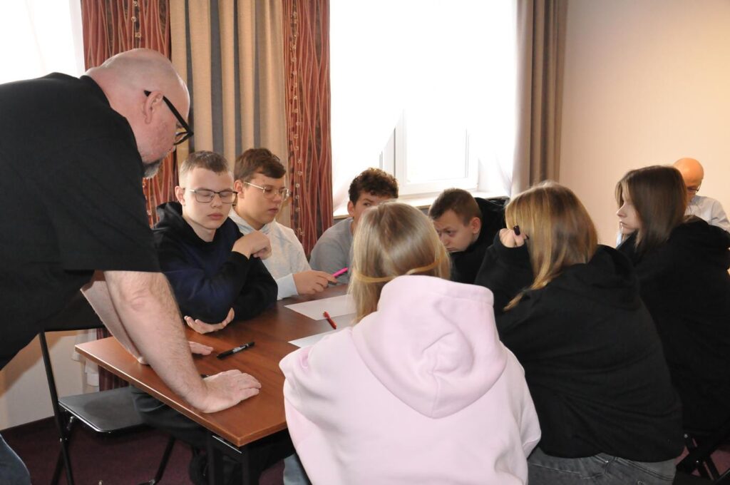 Mężczyzna z bródką i okularach stoi przy stole, przy którym siedzi młodzież.