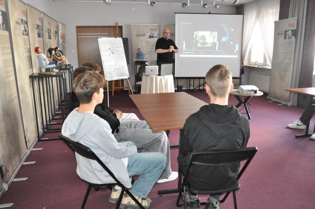 Przy stole siedzi kilka nastolatków. W dalszym planie stoi łysy mężczyzna z iwą bródką i okularach. Obok ekran z wyświetlonym kadrem filmu.
