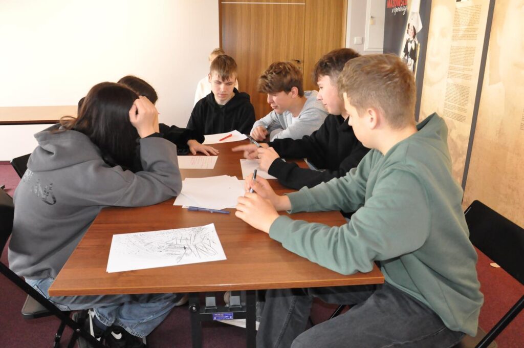 Grupa młodzieży siedzi przy stole. Notują coś na białych kartkach.