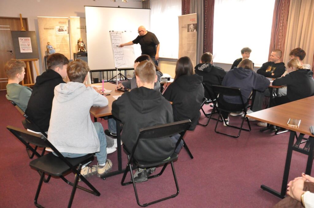 Grupa młodzieży siedzi przy dwóch stołach. W dalszym planie łysy mężczyzna w okularach rysuje na dużej białej planszy. Pomieszczenie ciepło oświetlone.