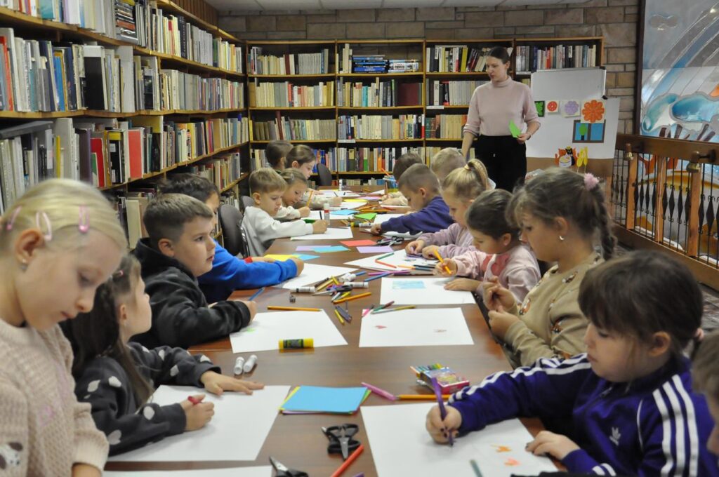 Grupa dzieci siedzi przy stole i maluje na kartkach. Wokół półki z książkami.