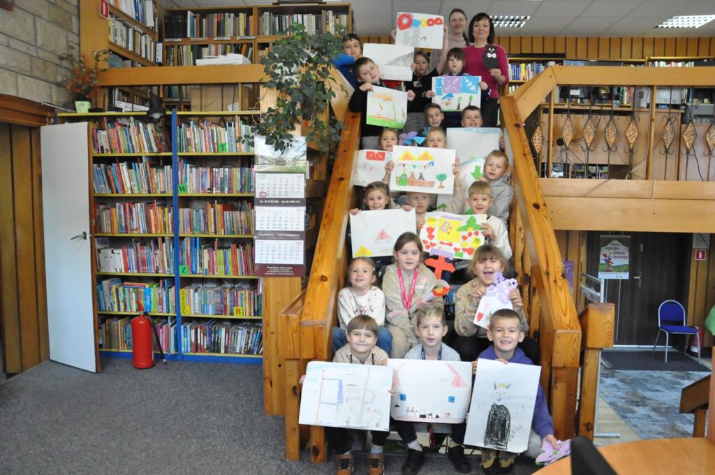 Grupa dzieci siedzi na schodach i pokazuje swoje prace plastyczne. Wokół Regały z książkami.