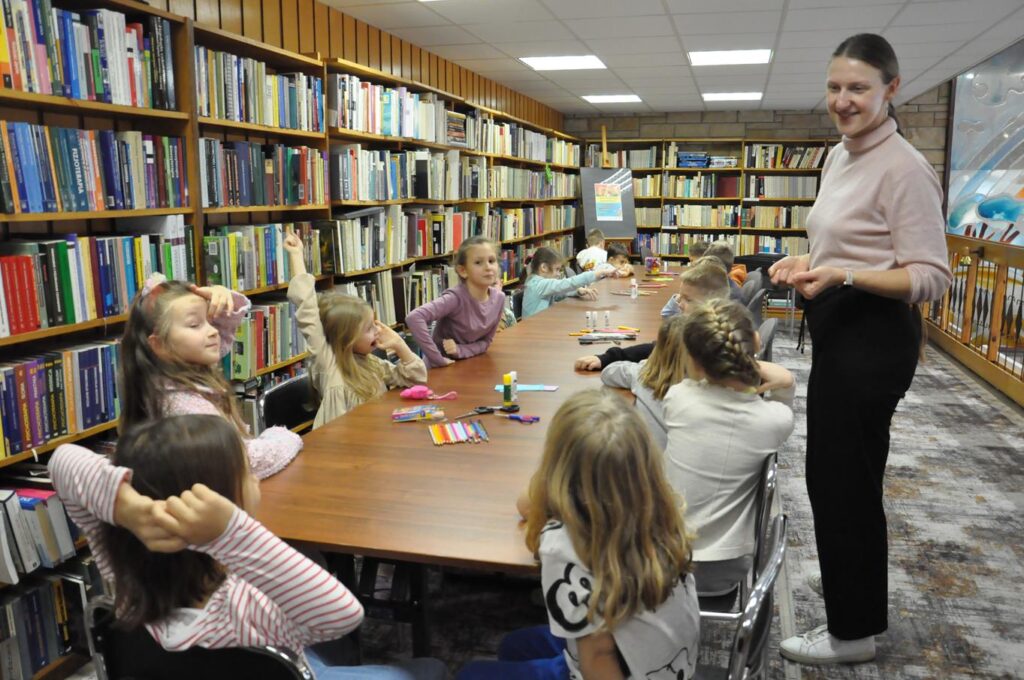 Grupa dzieci siedzi przy stole na którym leżą materiały plastyczne. Obok stoi kobieta w jasnym golfie. Wokół półki z książkami.