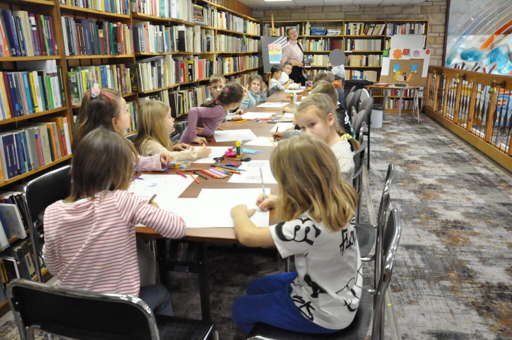 Grupa dzieci siedzi przy stole i maluje na białych kartkach. Wokół półki z książkami.