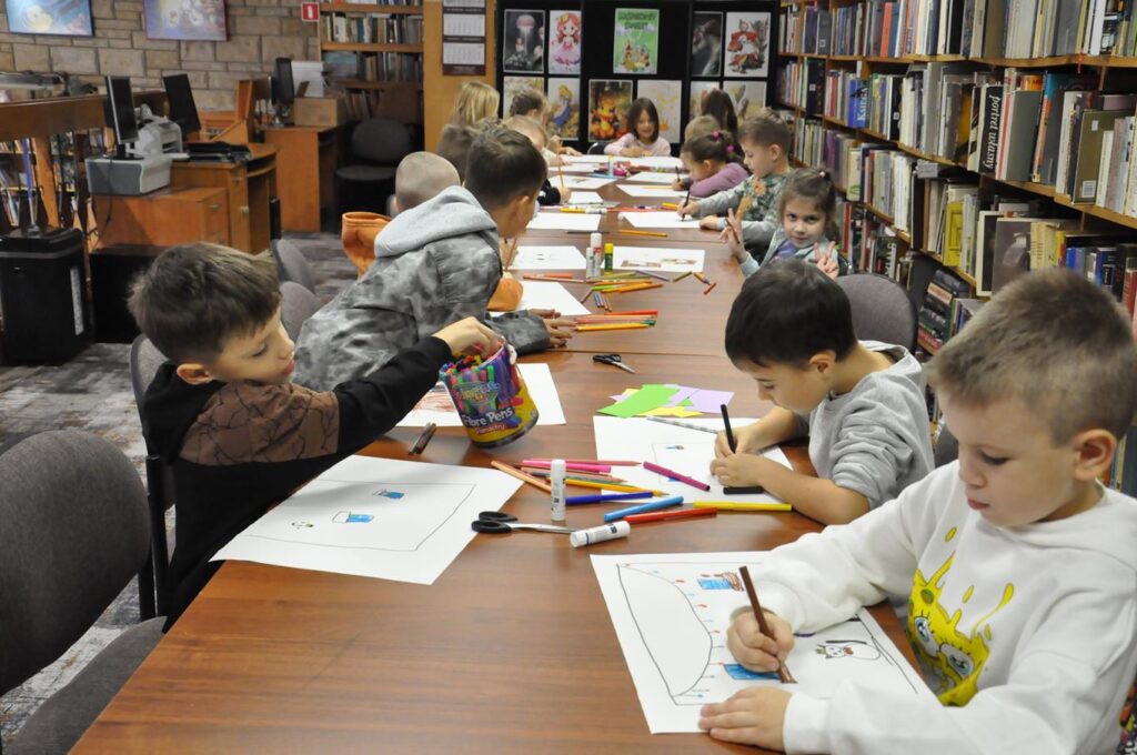 Grupa dzieci siedzi przy stole i maluje prace plastyczne. Wokół regały z książkami.