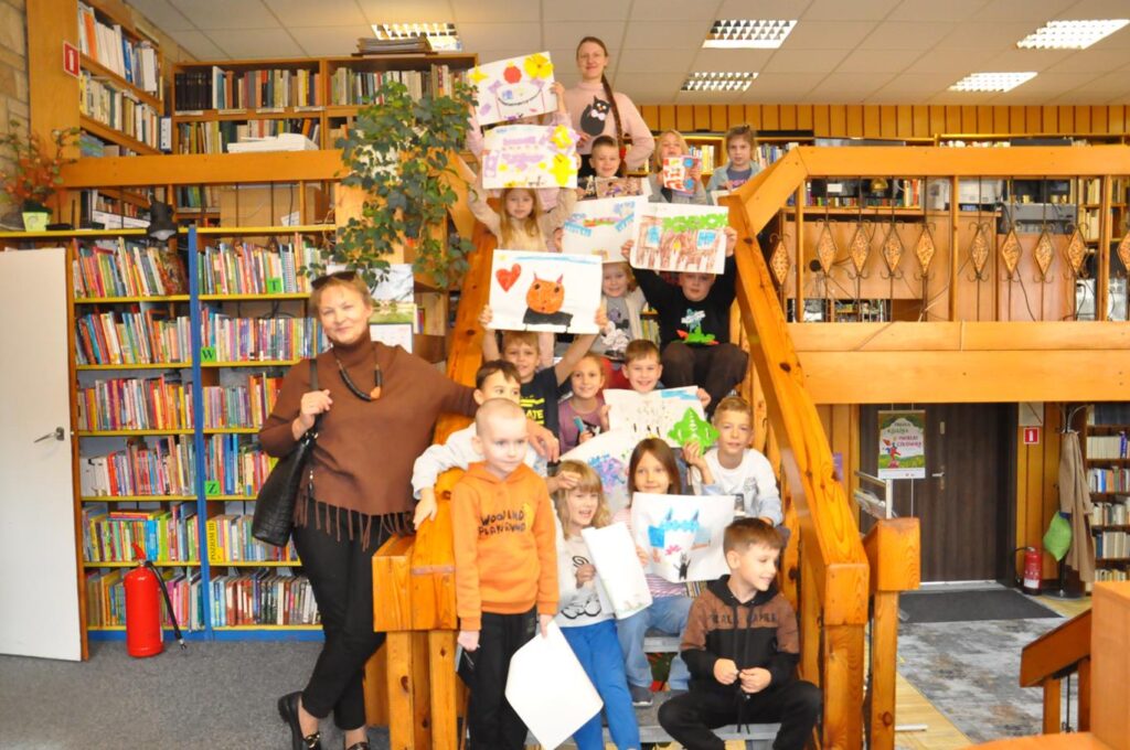 Duża grupa dzieci siedzi na schodach. Wszystkie eksponują swoje prace plastyczne. Wokół regały z książkami.