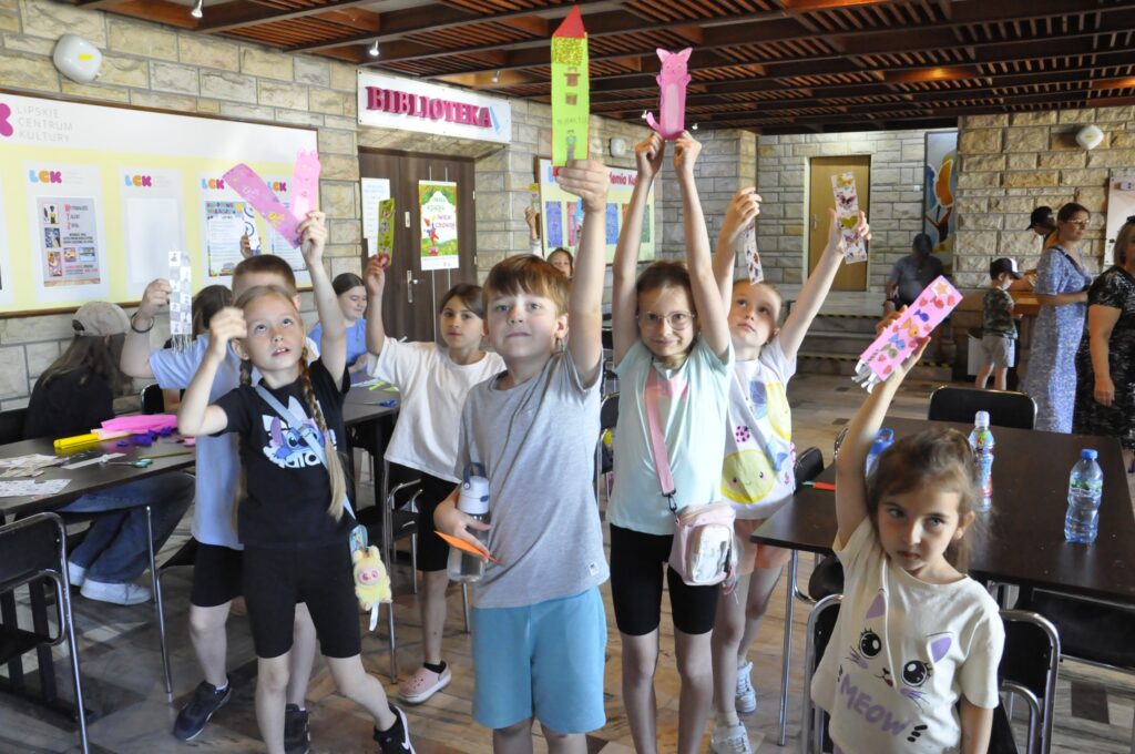 Dzieci w letnich ubraniach podnoszą kolorowe kartki papieru do góry. Pomieszczenie ciepło oświetlone.
