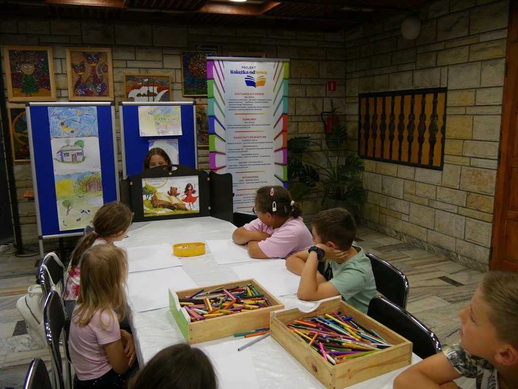 Dzieci siedzą przy stole i oglądają teatrzyk kamishibai w wykonaniu dziewczynki.