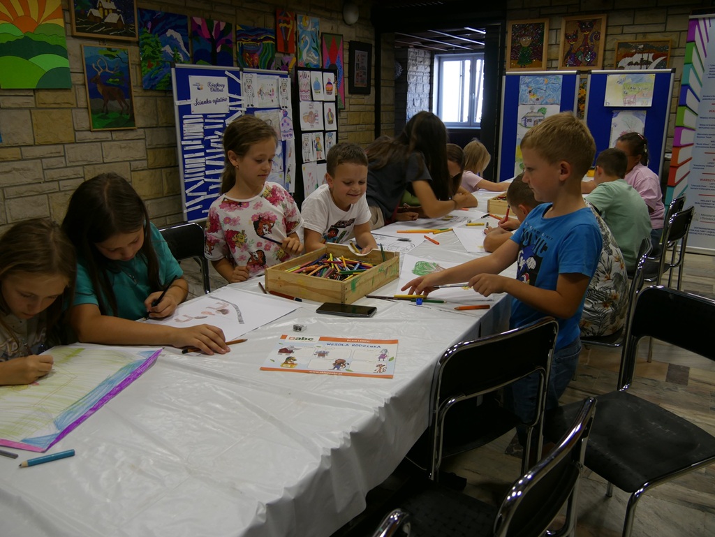 Dzieci w letnich ubraniach siedzą przy stole i wykonują prace plastyczne. Wokół niebieskie tablice z przypiętymi pracami.