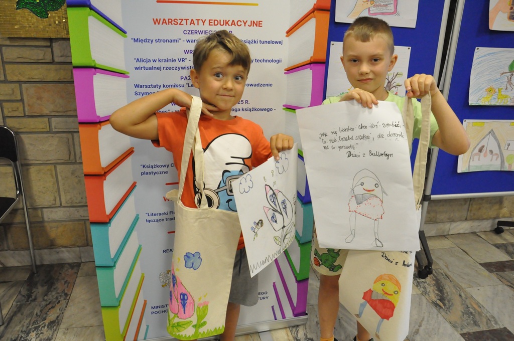 Dwaj chłopcy eksponują kolorowe torby i prace plastyczne. Za nimi kolorowy rollup.