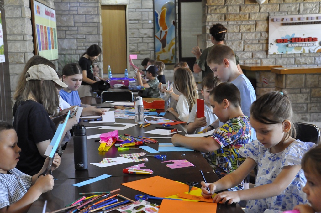 Dzieci w letnich ubraniach siedzą lub stoją przy stole i wykonują prace plastyczne.