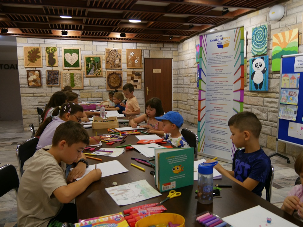 Dzieci w letnich ubraniach siedzą przy stole i wykonują prace plastyczne. Za nimi rollup z nazwą i spisem projektu. pomieszczenie ciepło oświetlone.