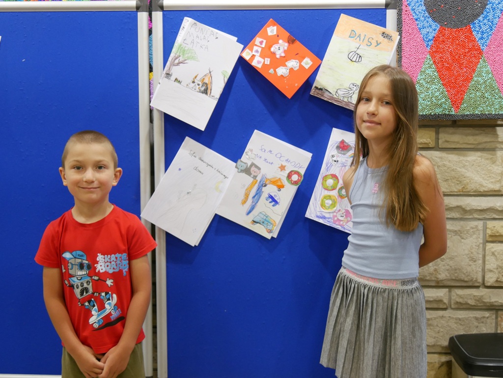 Chłopiec i dziewczynka stoją przy niebieskiej tablicy z pracami plastycznymi.