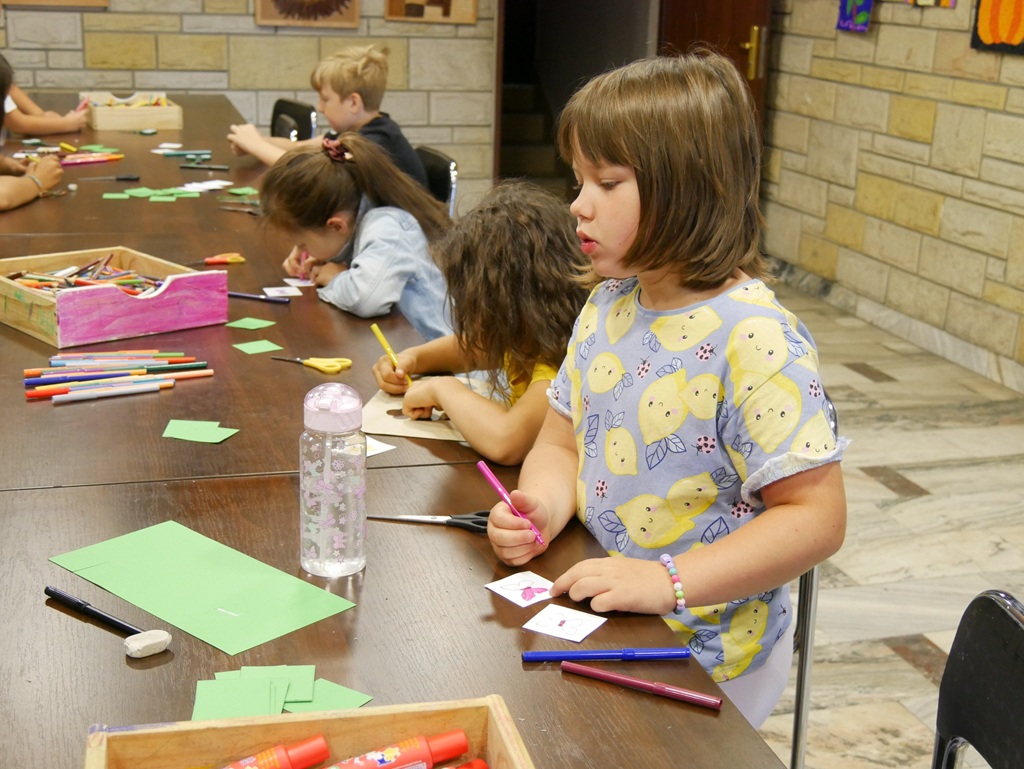 Dzieci siedzą przy stole i wykonują prace plastyczne. Pomieszczenie ciepło oświetlone.