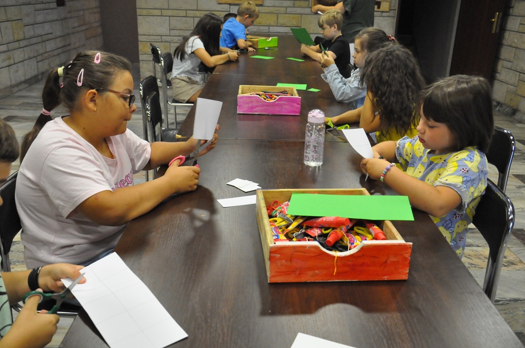 Dzieci w letnich ubraniach siedzą przy stole i wykonują prace plastyczne. Pomieszczenie ciepło oświetlone.