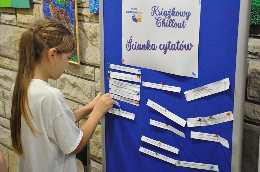 Dziewczynka przypina do niebieskiej tablicy karteczkę. Pomieszczenie ciepło oświetlone.