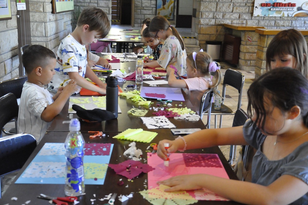 Dzieci w letnich ubraniach stoją lub siedzą przy stole i wykonują prace plastyczne. pomieszczenie ciepło oświetlone.