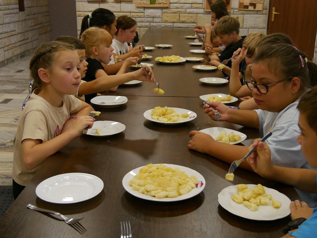 Dzieci siedzą przy stole i jedzą kopytka. Pomieszczenie ciepło oświetlone.